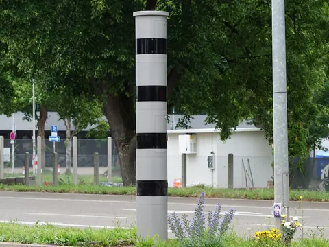 A grey speed camera column with black bands beside a town road