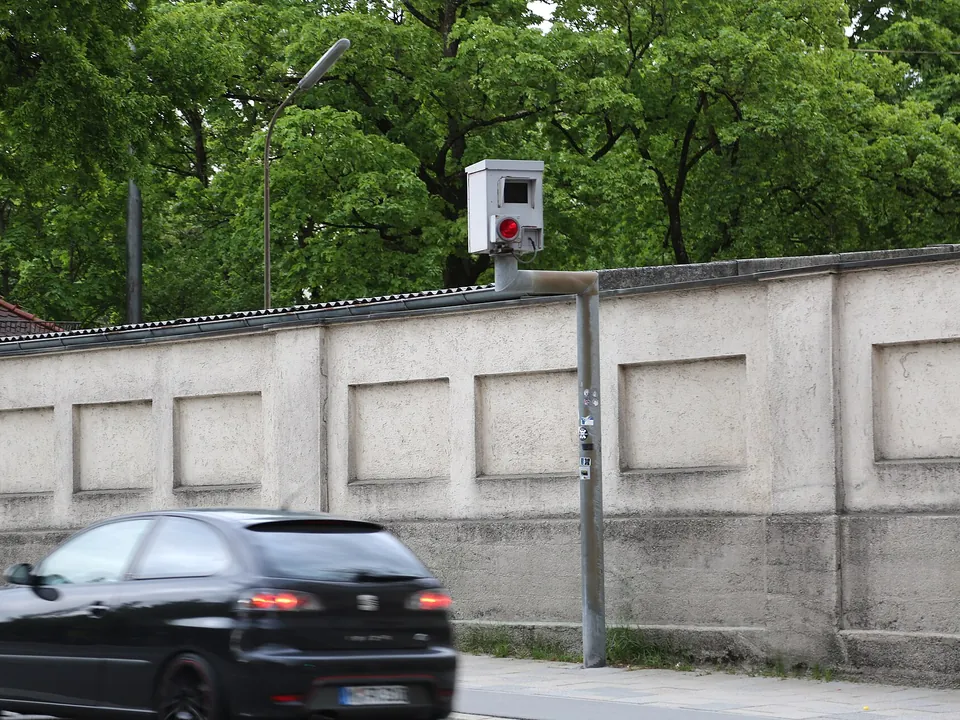 A red light camera box on a bracket above a road as a car passes