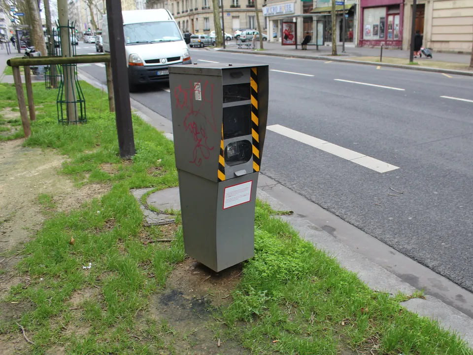A grey speed camera box with yellow and black stripes on a post beside a Paris avenue