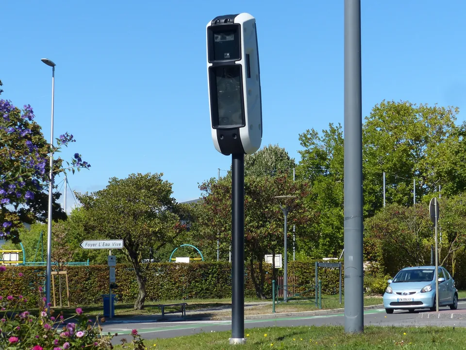 A slim speed camera on top of a tall black post beside a town road