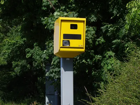 A yellow speed camera on a grey pole beside a road verge