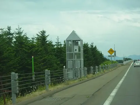 A tall Orbis speed camera housing behind wire mesh beside a highway